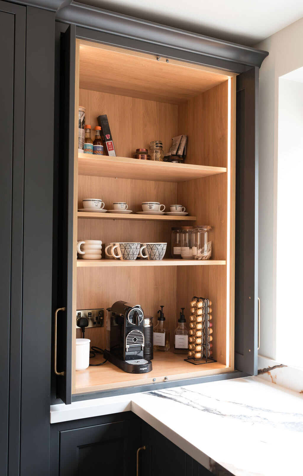 A built-in kitchen cabinet with black exterior and wood interior, illuminated by built-in lighting, displays shelves with mugs, glasses, spices, and coffee supplies. The counter features a coffee machine, capsule holder, and various bottles.