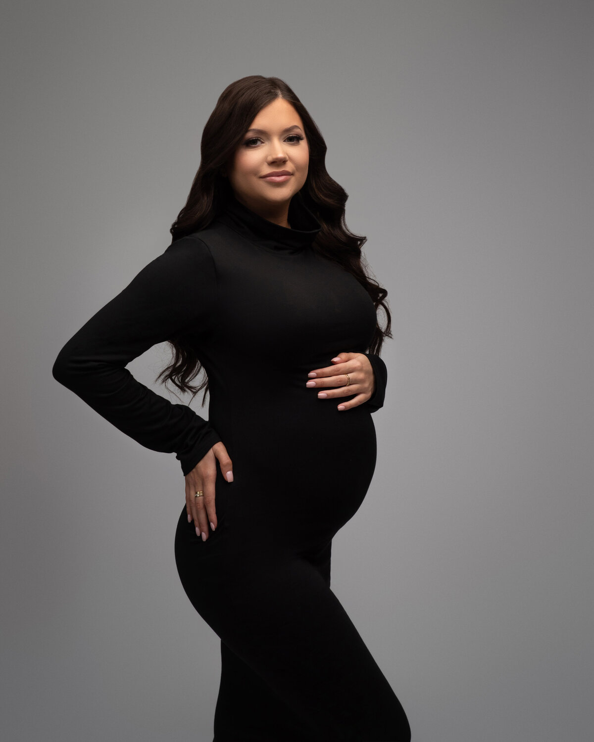 pregnant woman with long brown hair posing with hand on back and the other hand on belly bump wearing turtle neck black dress