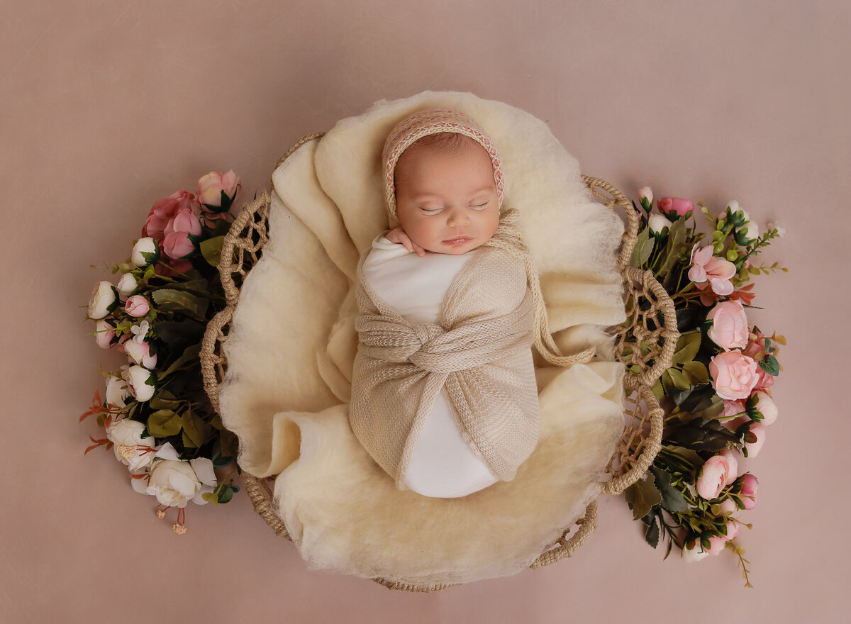 Brooklyn newborn photographer captures baby girl for a newborn photoshoot. Aerial image. She is wrapped in cream swaddle and placed in a basket atop of fresh flowers. She is sleeping. Captured by top Brooklyn newborn photographer Chaya Bornstein