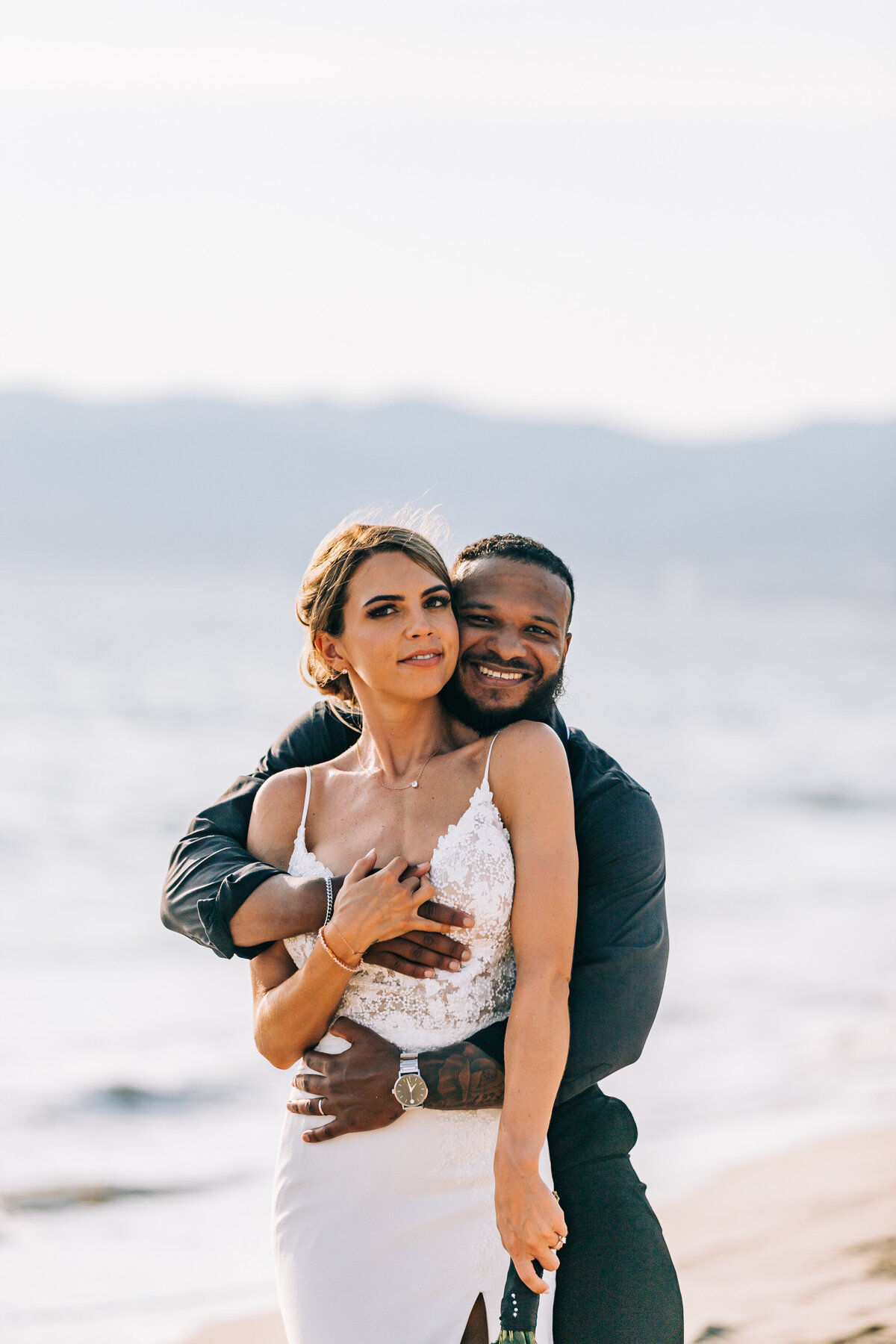 A groom embraces his bride from behind as they stand on a beach. The bride is wearing a white dress with lace detailing, and the groom is dressed in dark attire. The ocean and mountains are visible in the background on a sunny day