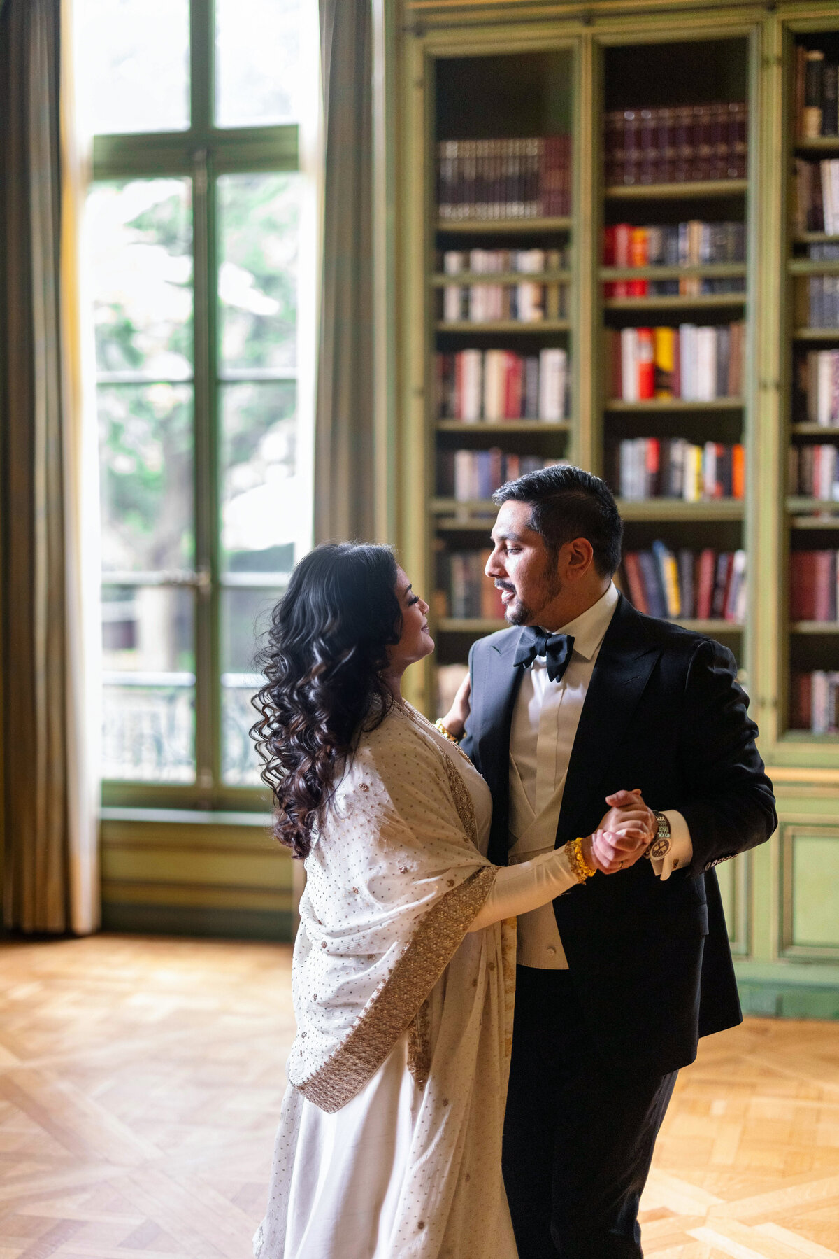 A couple dances in a luxurious room with a large window and floor-to-ceiling bookshelves. The man is wearing a tuxedo, and the woman is in a traditional outfit. Sunlight filters through the window, creating a warm atmosphere.