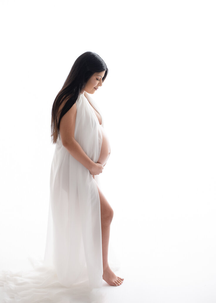 Maternity photography -- Pregnant woman in flowing white gown stands in profile against a bright white background in a Birmingham maternity photography studio