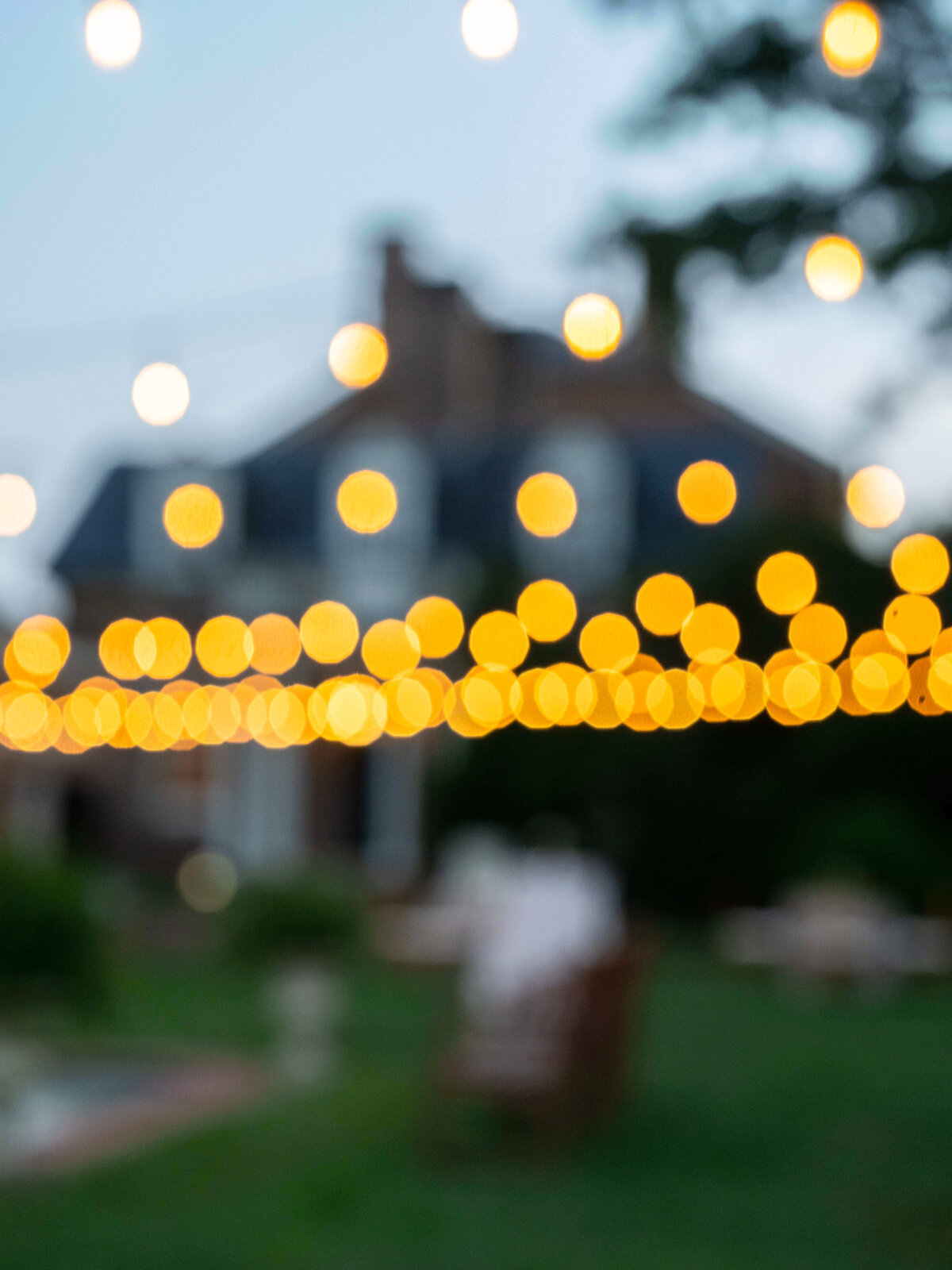 Blurred image of a house with numerous string lights creating a warm, glowing effect. The focus is on the bokeh effect of the lights against a dim, twilight sky. The foreground shows an out-of-focus garden area.