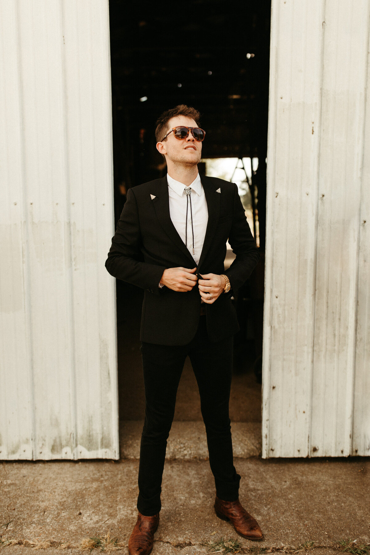 Boho groom with bolo tie, black suit and sunglasses getting ready in front of a barn on the morning of his wedding