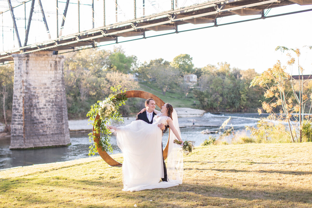 groom carries bride from alter with Faust Bridge behind at Milltown Historic District in New Braunfels sunset
