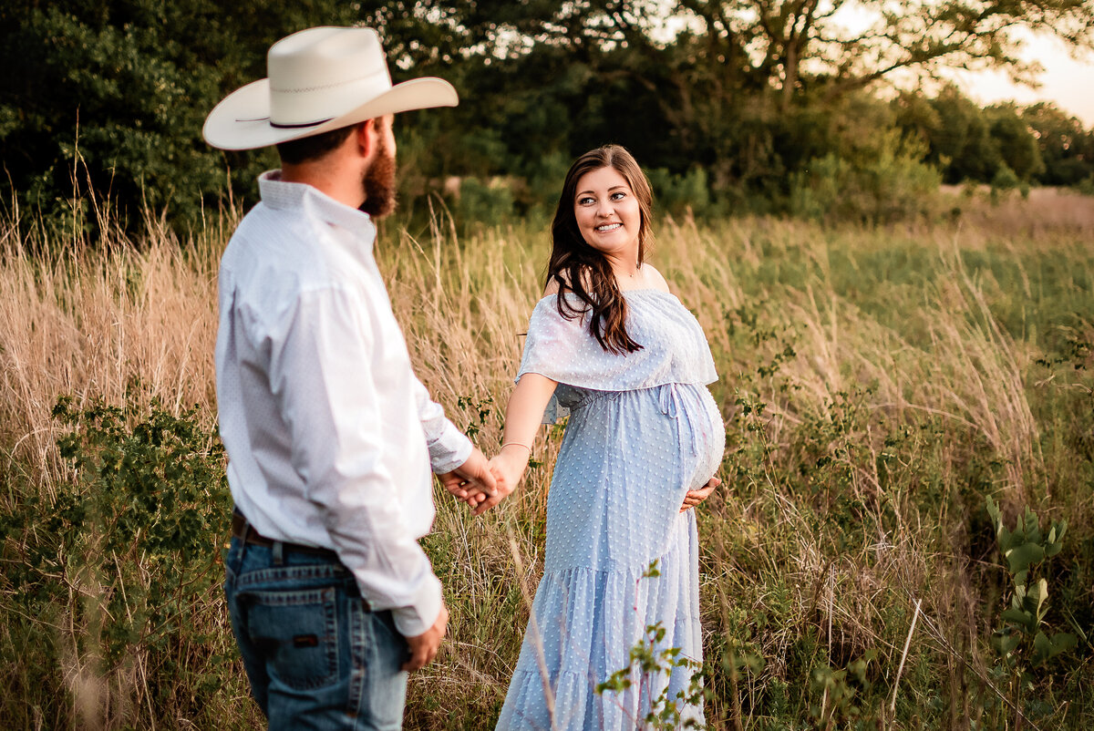 A pregnant woman wearing a blue dress holds her husband's hand as she leads him through a field at Brazos Bend State Park.