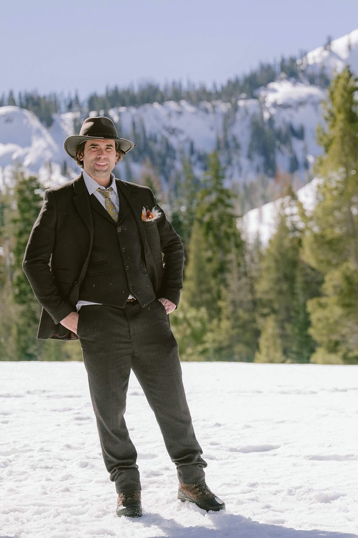Groom in brown 3 piece suit and hat posing after Palisades Tahoe elopement snowy mountain in background