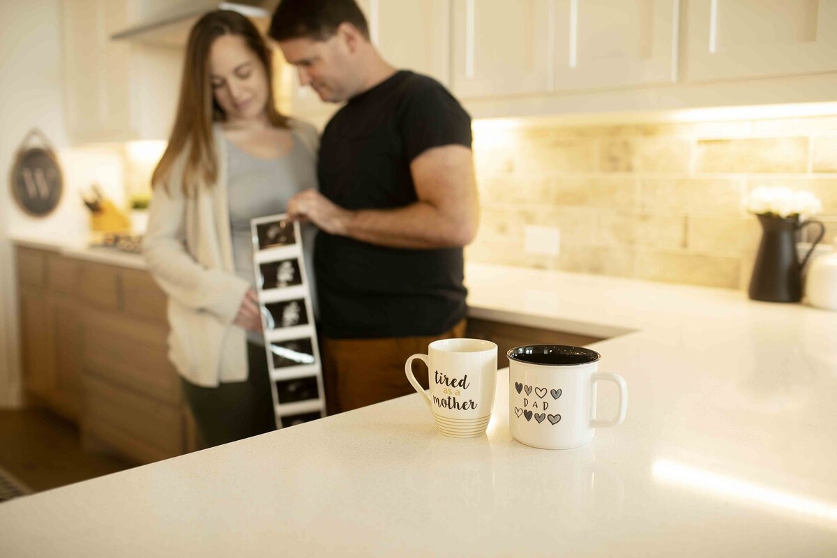 A casual maternity photo of mom in a tank top, off-the-shoulder sweater, and leggings, standing in the kitchen and cradling her baby bump, with soft natural light coming through the window, captured by Chunky Monkey Photography.