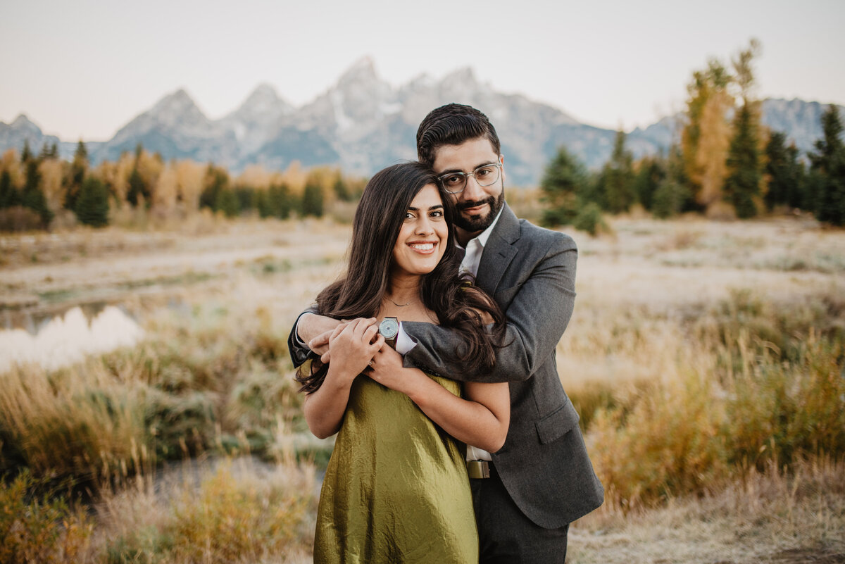 man holding his arms around his girlfriends shoulders as she reaches up to hold his hands as they both smile for their fall engagement session in the grand tetons with photographers in jackson hole