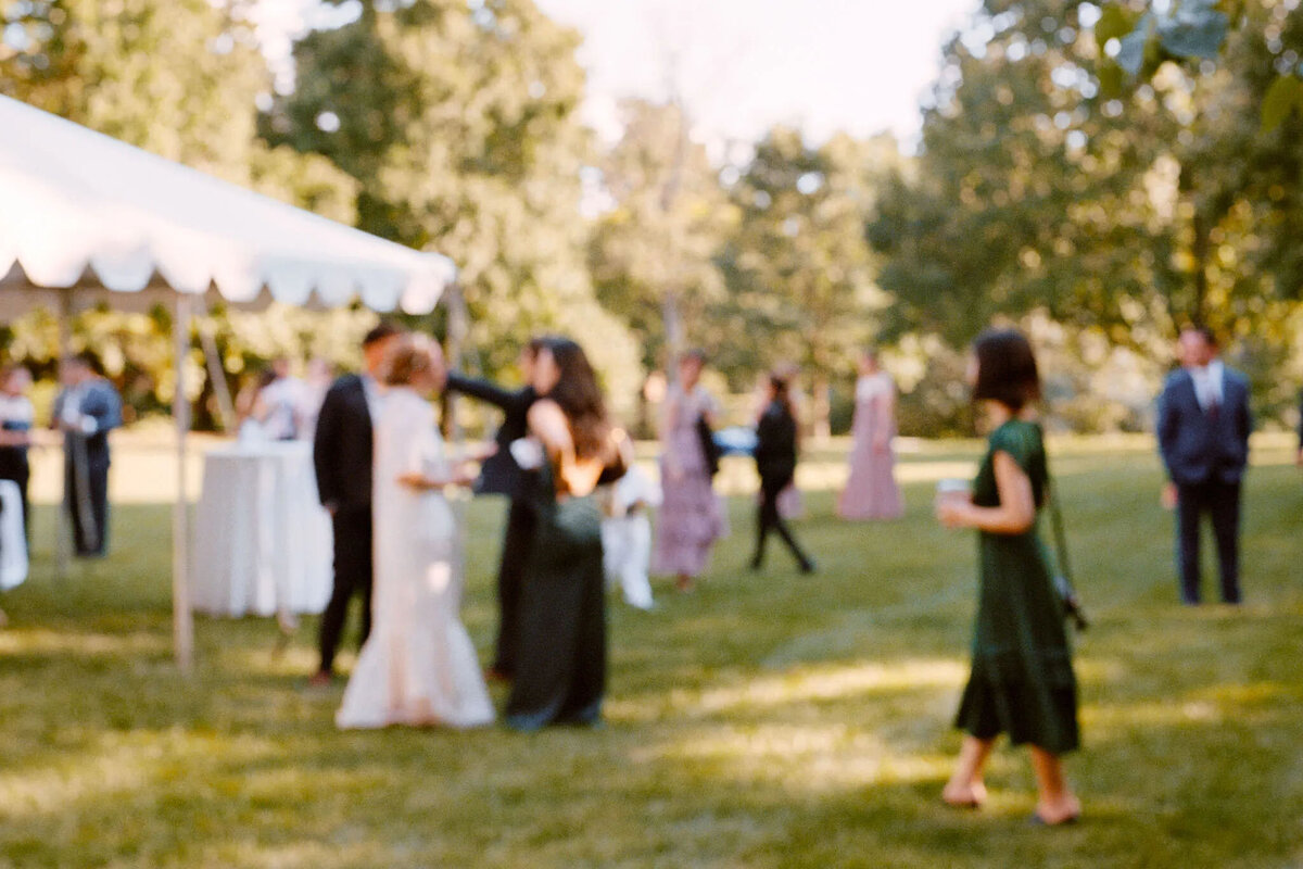 A group of people in formal attire mingle outdoors on a grassy lawn with trees and a white tent in the background; the image, captured by an NJ wedding photographer, is out of focus.