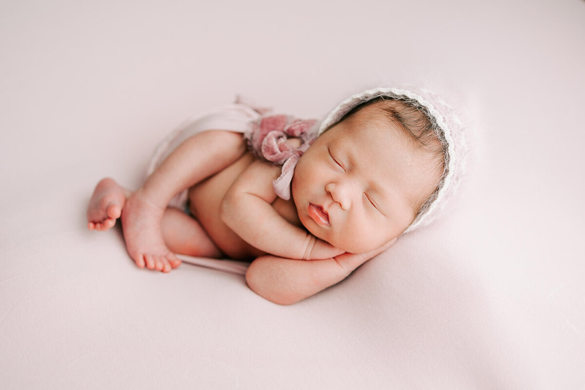 girl wearing bonnet with hands under her chin sleeping on pink backdrop