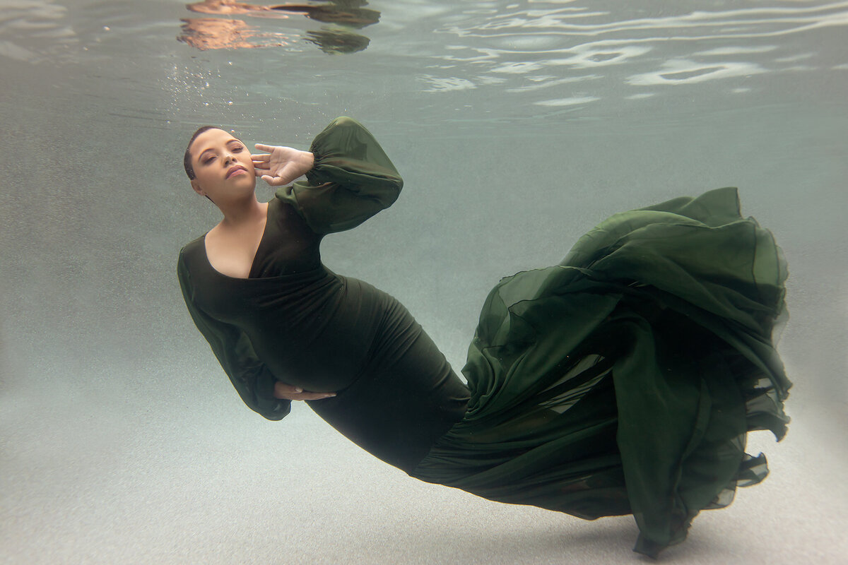 A serene underwater portrait showcasing a woman gracefully posed in a flowing dark green gown. The fabric swirls around her, creating soft, organic shapes against the water’s clarity, enhancing the peaceful ambiance.