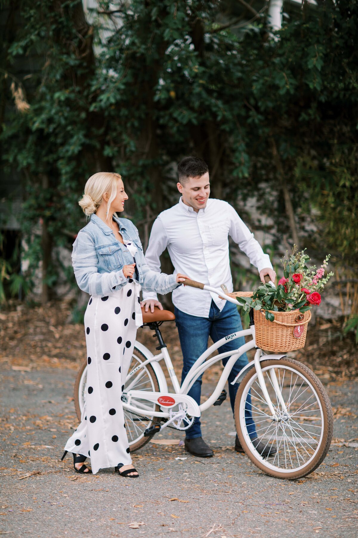 Young blonde girl in polka dot outfit walks beside a handsome guy with a white bike and flowers in the basket for their engagement session with Danielle Defayette Photography in Charleston