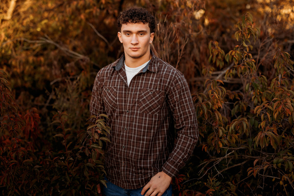 High school teen boy stands with hands in pockets in front of fall bushes
