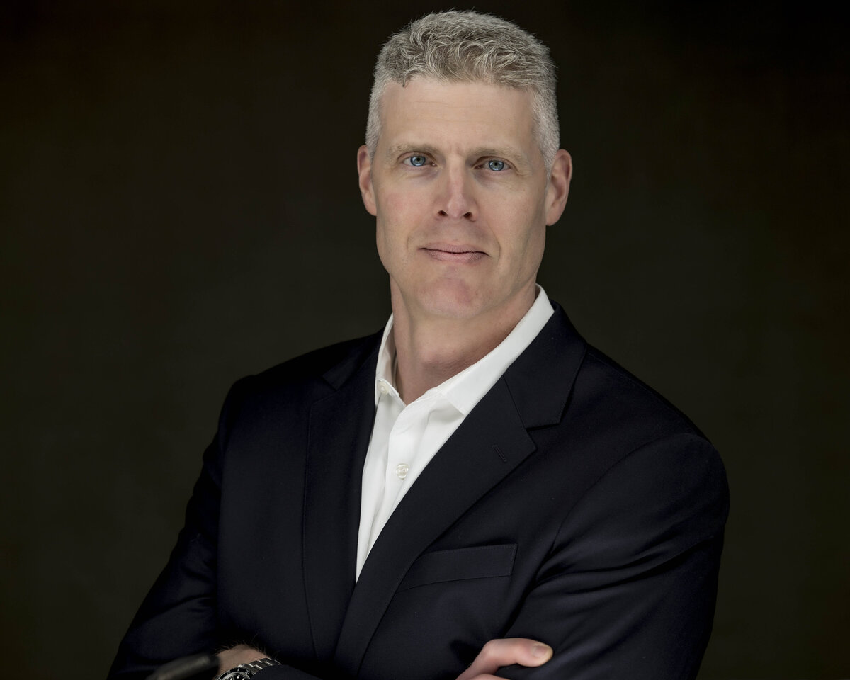 Business headshot in MA of man in dark suit jacket and open-collar white shirt against dark background by Rob Faber Photography.