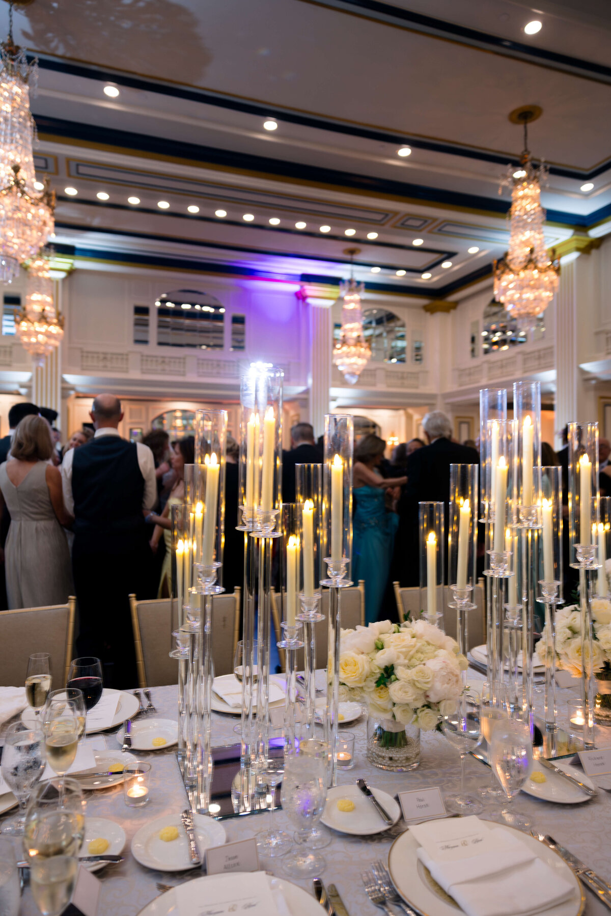 A luxurious event hall is elegantly decorated with tall candle centerpieces and white floral arrangements on tables. Guests in formal attire mingle beneath ornate chandeliers, with soft lighting creating a sophisticated atmosphere.