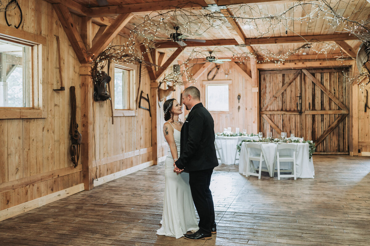 A newlywed couple stands intimately, holding hands and gazing at each other, in the rustic wooden interior of The Gentry Farm's barn in Danville, Virginia, adorned with string lights, captured by Quinn Photography LLC.