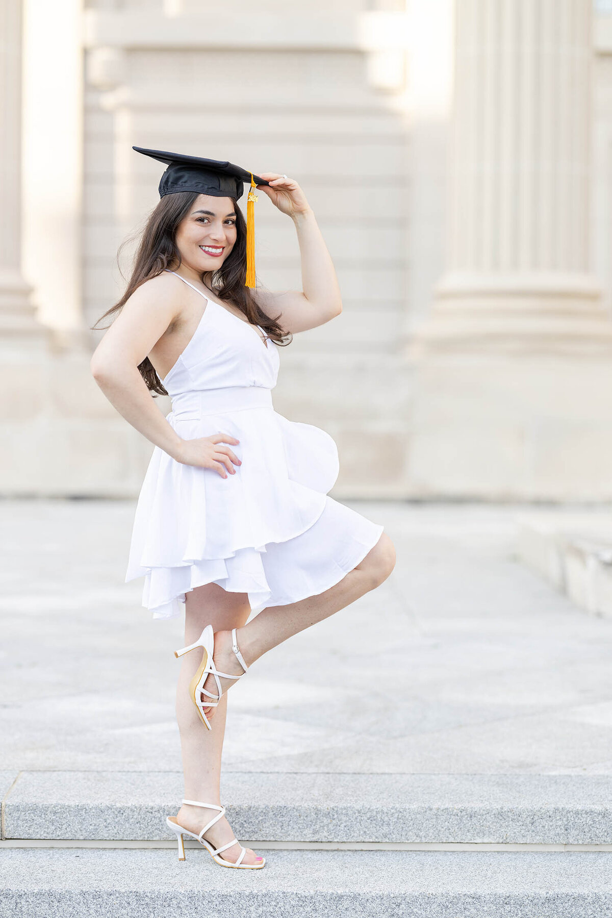 Graduate girl wearing a knee length white dress has one knee bent as she holds her cap with one hand smiling during her graduate photo session