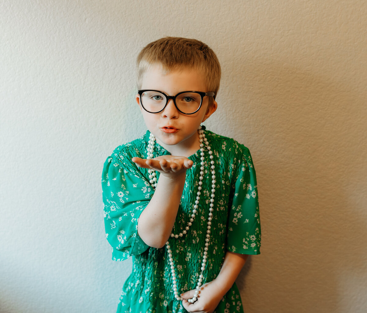 A little boy stands in a green dress, wearing a pearl necklace and black glasses, and blows a kiss at the camera