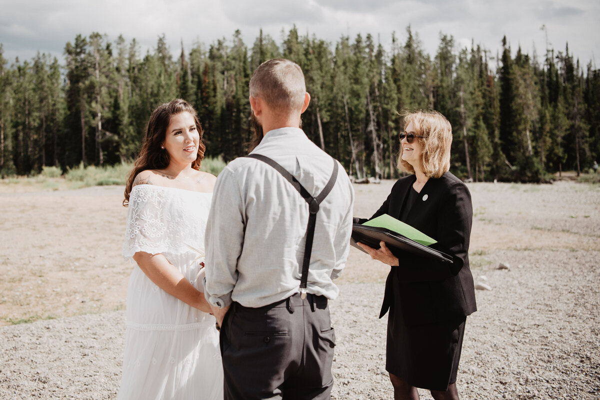 beach wedding in the grand tetons with couples getting married on the sand next to the woods as they hold hands and their officiant shares kind words for a jackson hole wedding