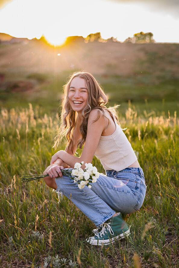 Girl in tank and jeans squats while holding a bouquet of flowers with sun behind her.