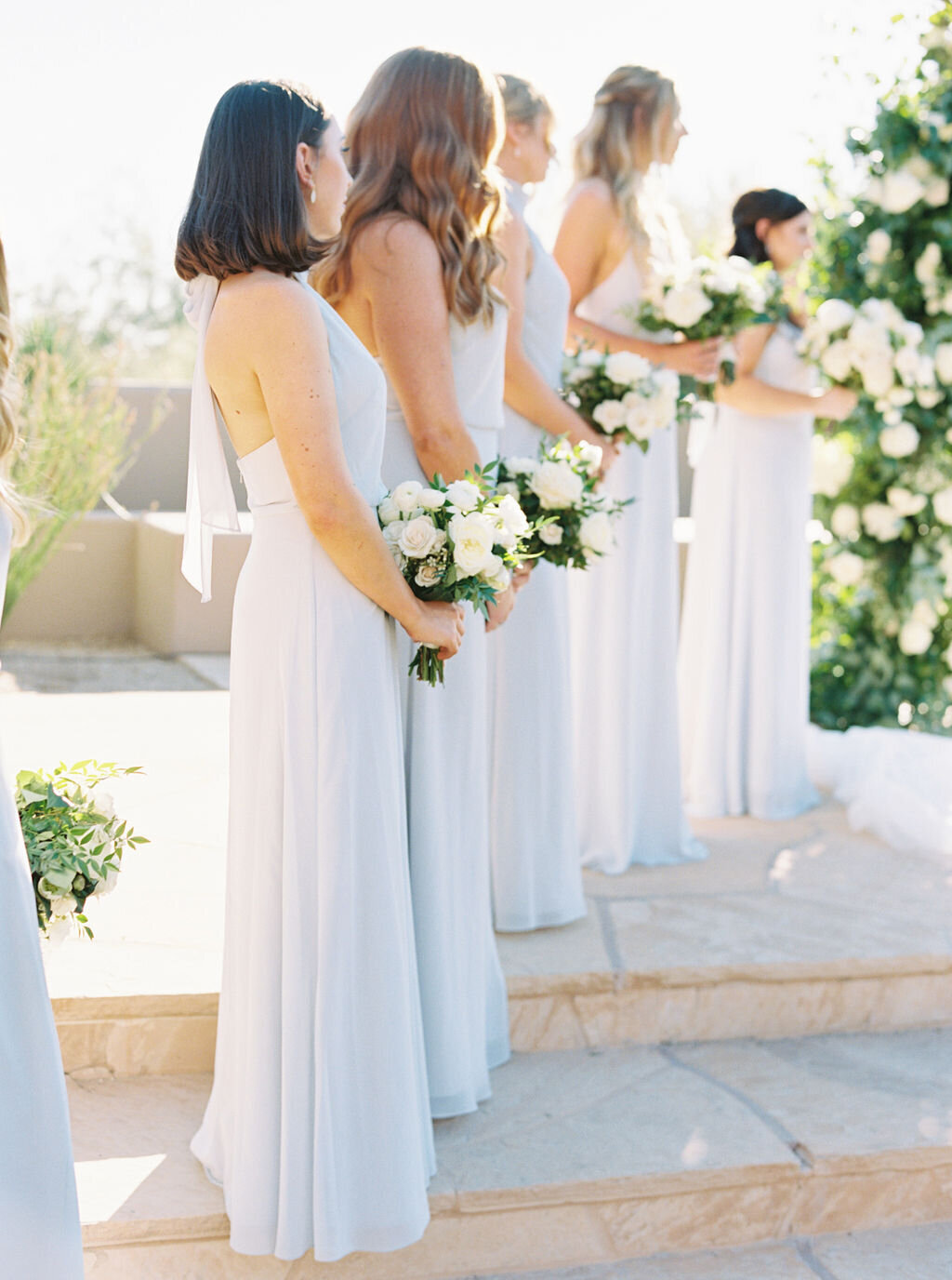 A beautiful lineup of bridesmaids in flowing blue dresses, holding classic white floral bouquets. This elegant wedding aesthetic perfectly complements a timeless and romantic ceremony setting.