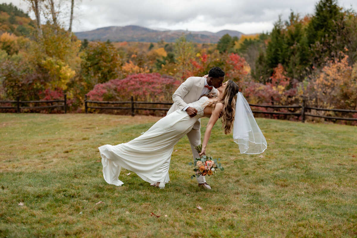 In a picturesque New England setting, a bride and groom share a romantic dance dip in a grassy field. The bride holds a bouquet and wears a veil, while the groom is in a beige suit. The scene, captured by an expert in elopement photography, features colorful autumn foliage and distant mountains.