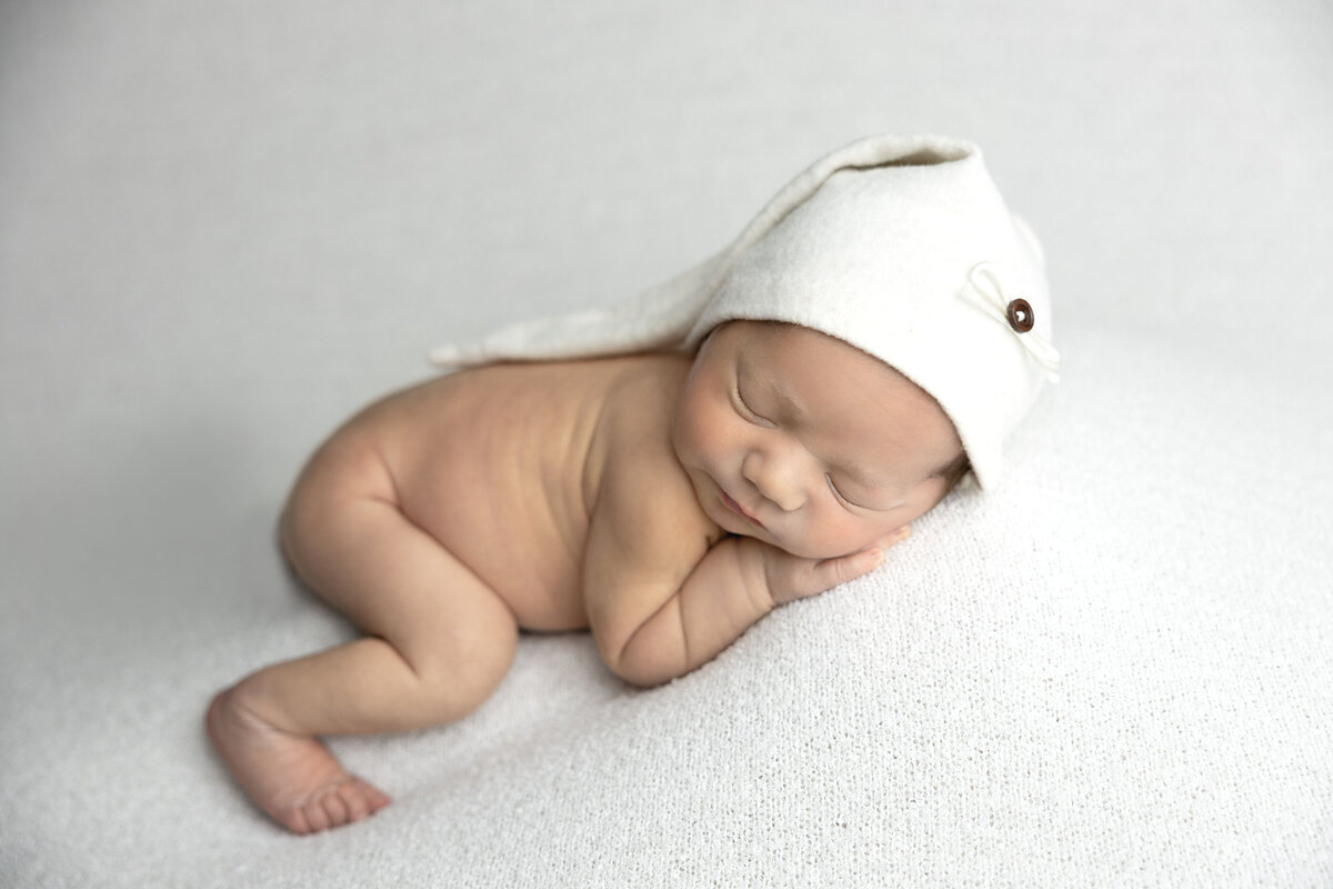 Baby boy sleeps on his side with a white sleeper hat on.