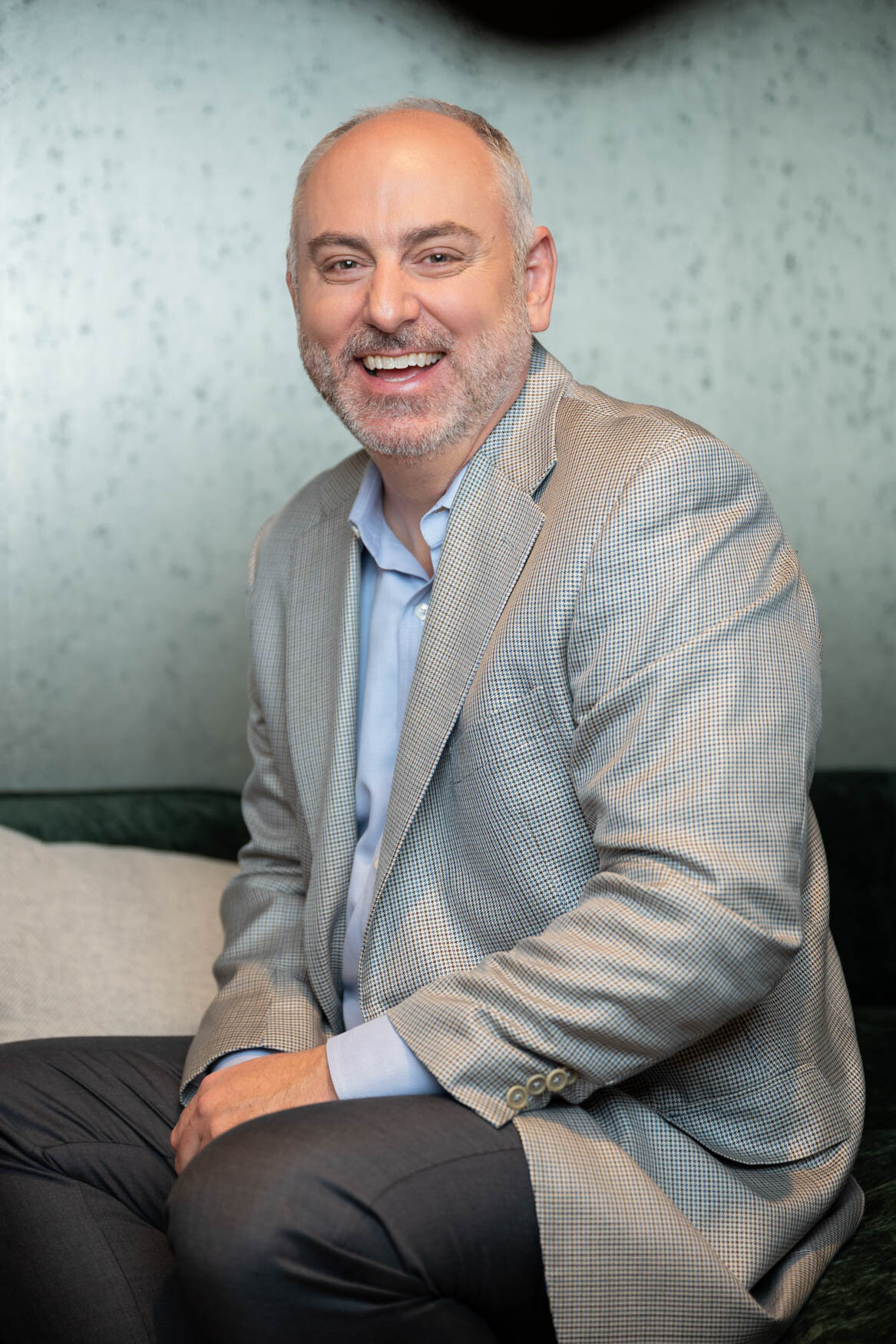 A portrait of a man laughing big to the camera. He is sitting on a green couch and is wearing a grey jacket and dark pants