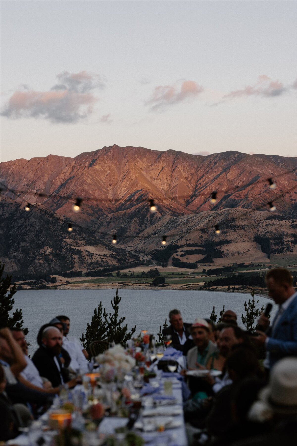 Guests seated at a long alfresco dinner table, listening to a speech as warm sunset light hits the mountains and lake behind them, with string lights glowing overhead.