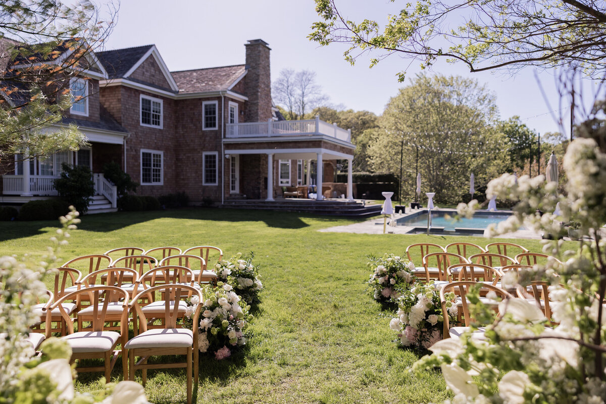 Elegant outdoor ceremony setup at a private Hamptons estate, featuring wooden chairs, lush floral aisle arrangements, and a view of the home and pool in the background.