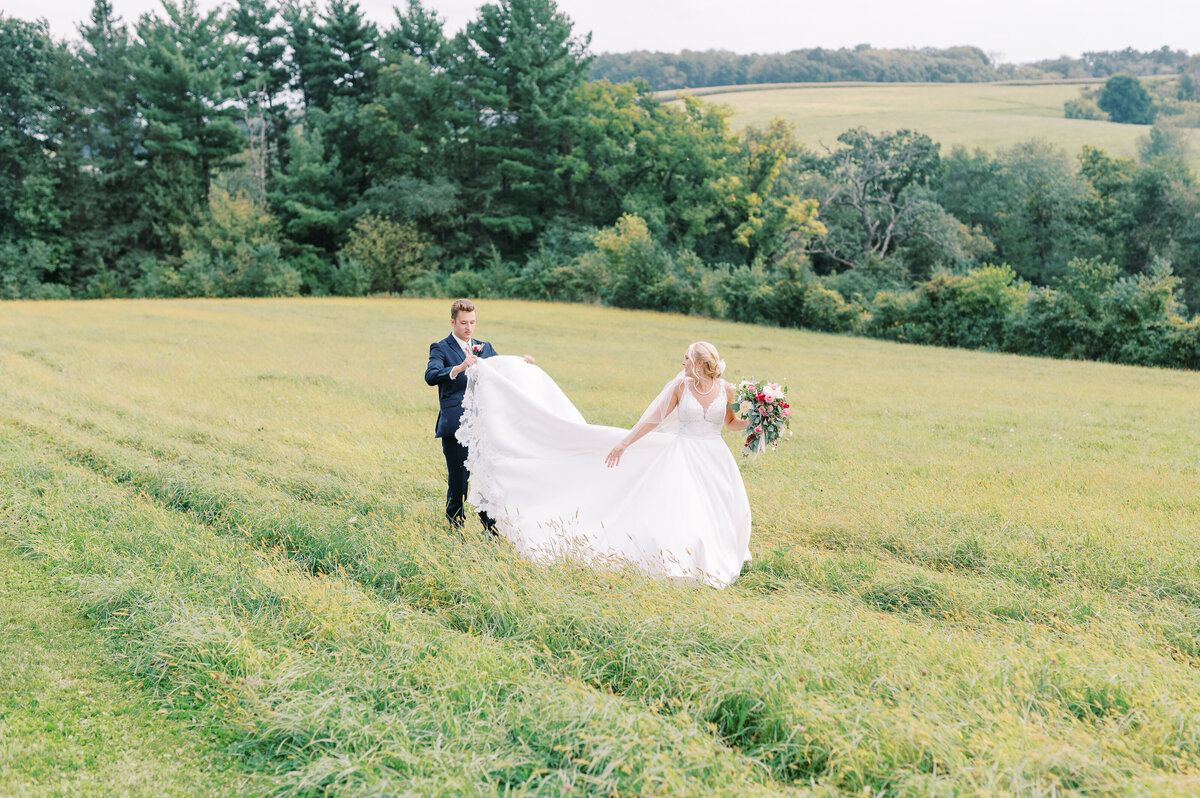 Groom carrying bride's train in a grassy field