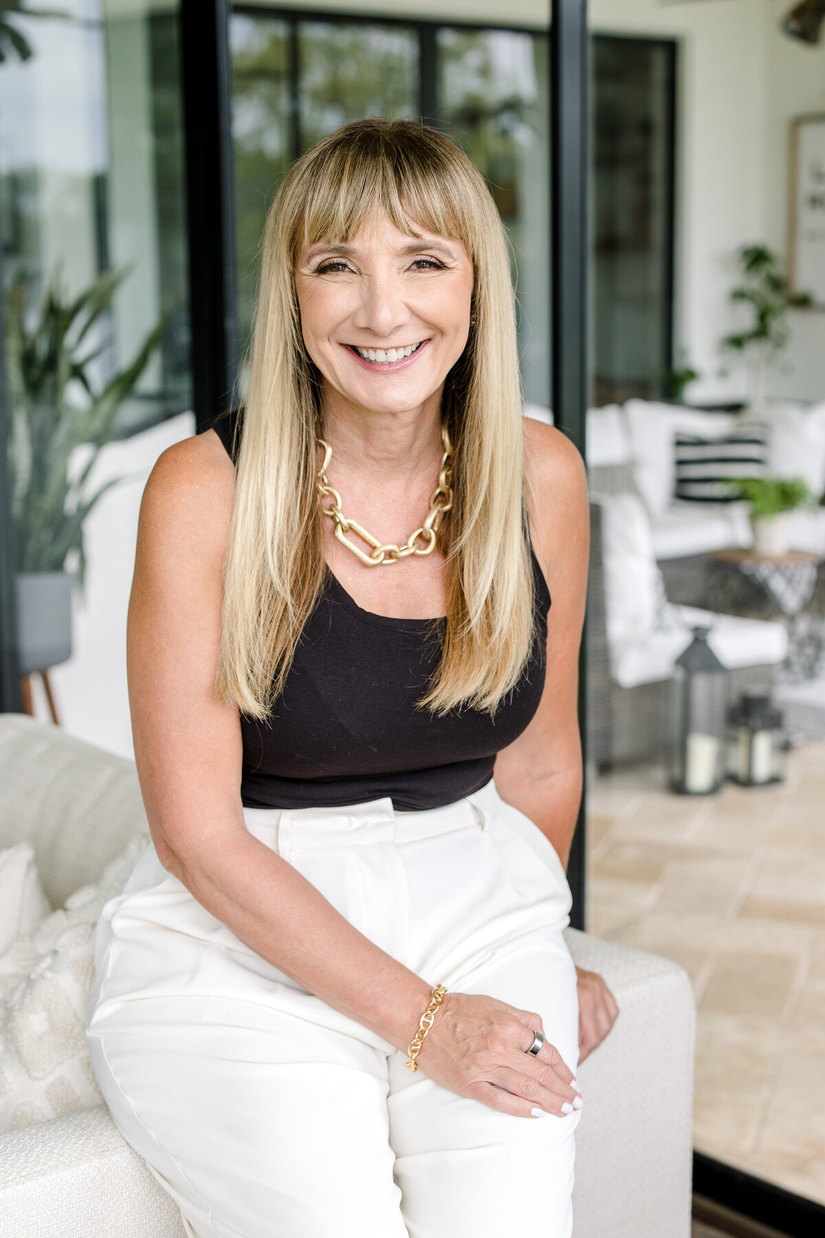 Business headshot of women with long blonde hair and a black shirt and white pants sitting on the edge of the couch cleaning one arm back and smiling on the porch captured by brand photographer near me