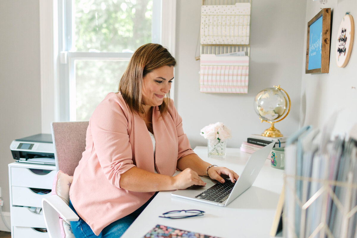 A woman in a pink blazer sits at a white desk, working on her laptop in a bright and organized home office. A gold globe, pink flowers, and neatly arranged files decorate the space.