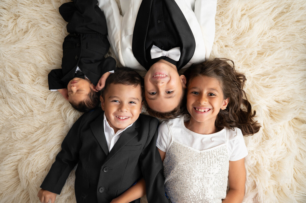 Three siblings in black and white formal outfits laying beside newborn baby.