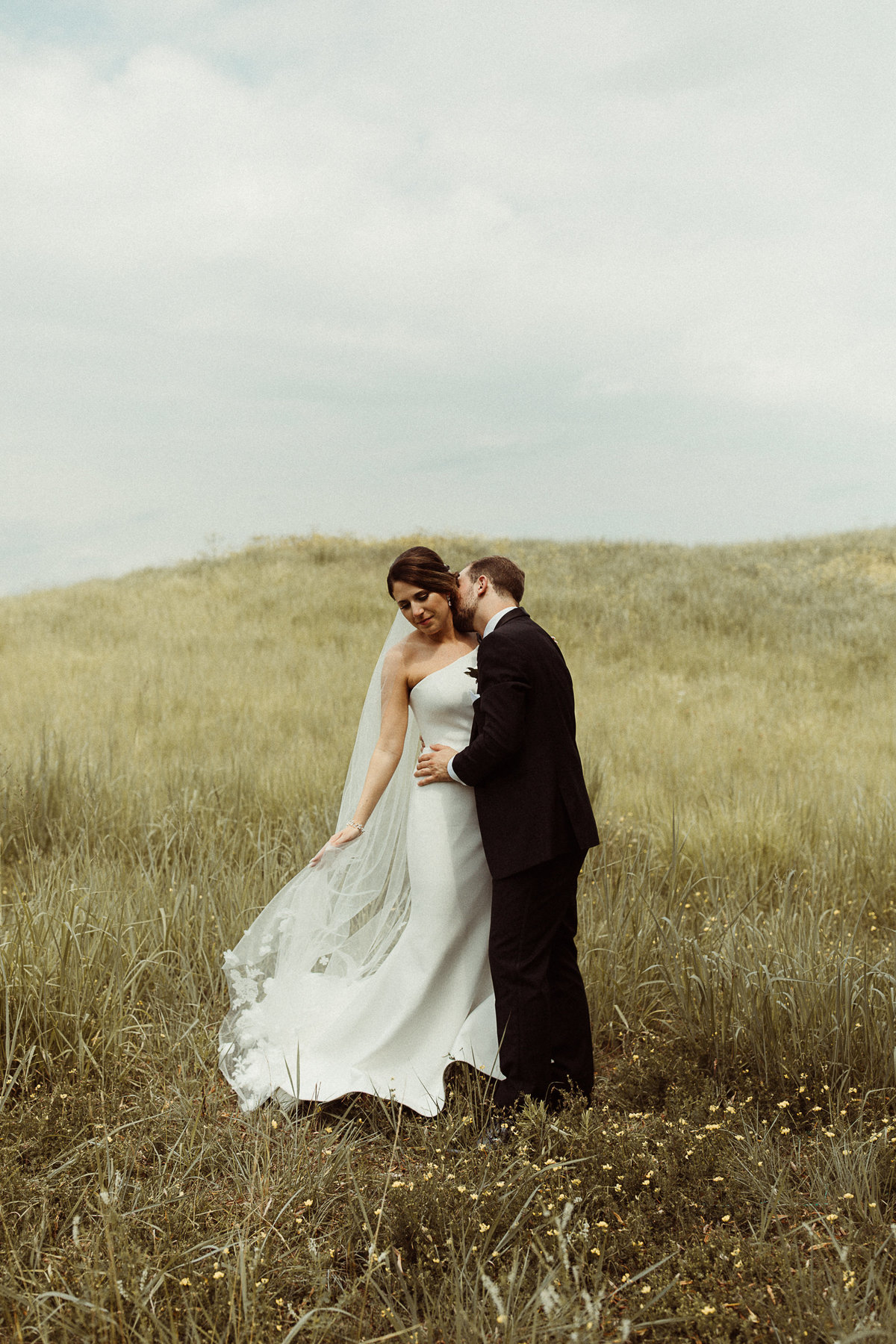 bride wearing asymmetrical dress in a field with the groom kissing her shoulder