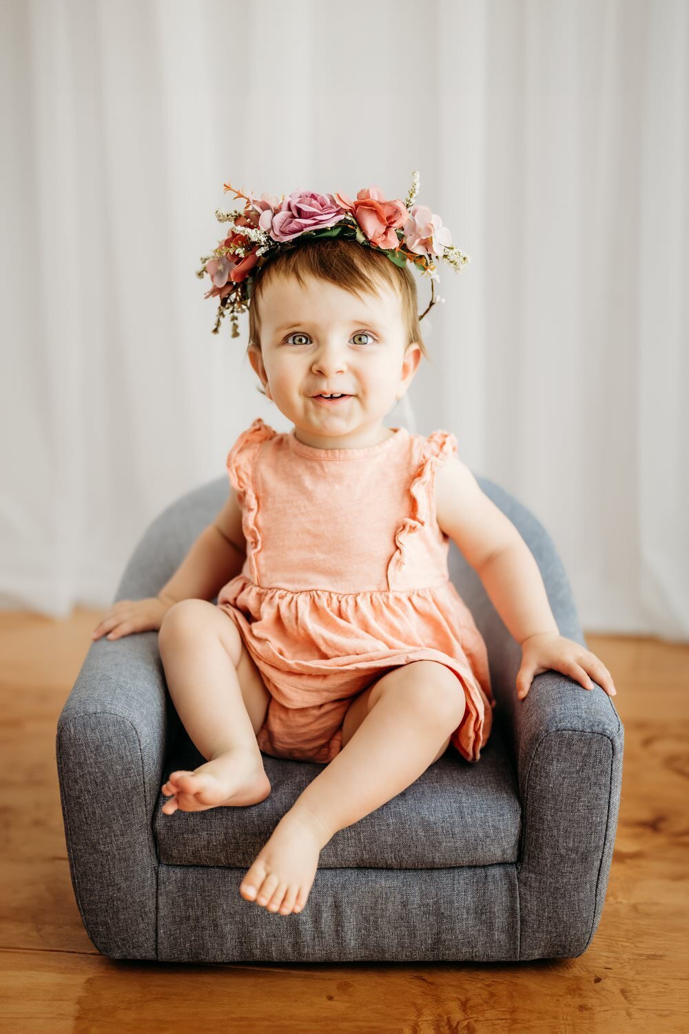 first birthday photo of baby girl. she smiles at camera and sits in a grey chair