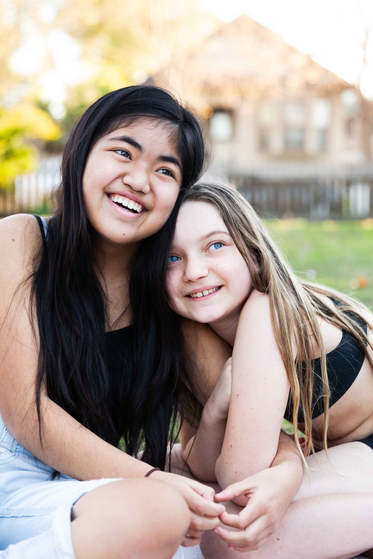 Sibling teenage sisters leaning sweetly on each other in the front yard of their home.