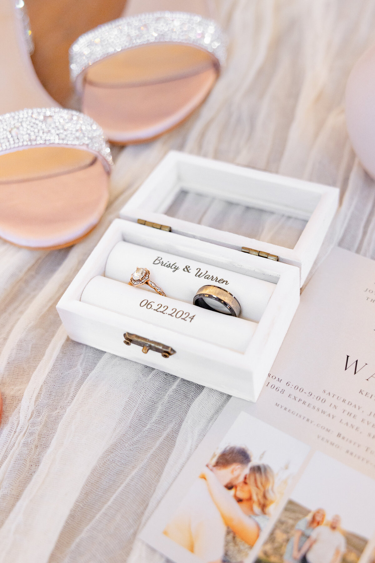 A beautifully captured wedding ring box featuring an engagement ring and a groom's band, engraved with the names "Bristy & Warren" and the wedding date "06.22.2024." Delicate details of bridal shoes and an invitation complement the scene. Photographed at Maplewood Events in Spanish Fork, Utah by Cassie Lee Photography.