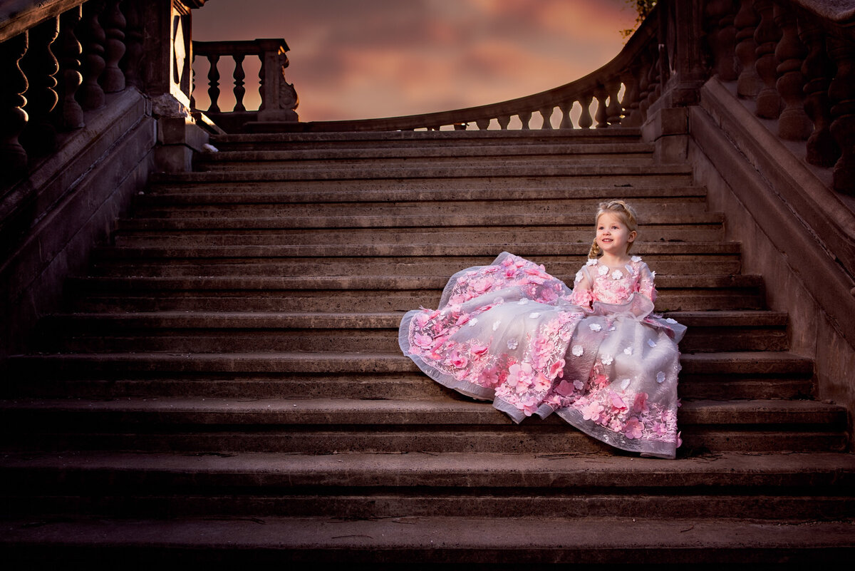 Regal portrait of a young girl in a flowing pink gown, seated on grand stone stairs at dusk. Captured in Leawood, Kansas, this dramatic and elegant setup is perfect for families seeking storybook-themed child photography.