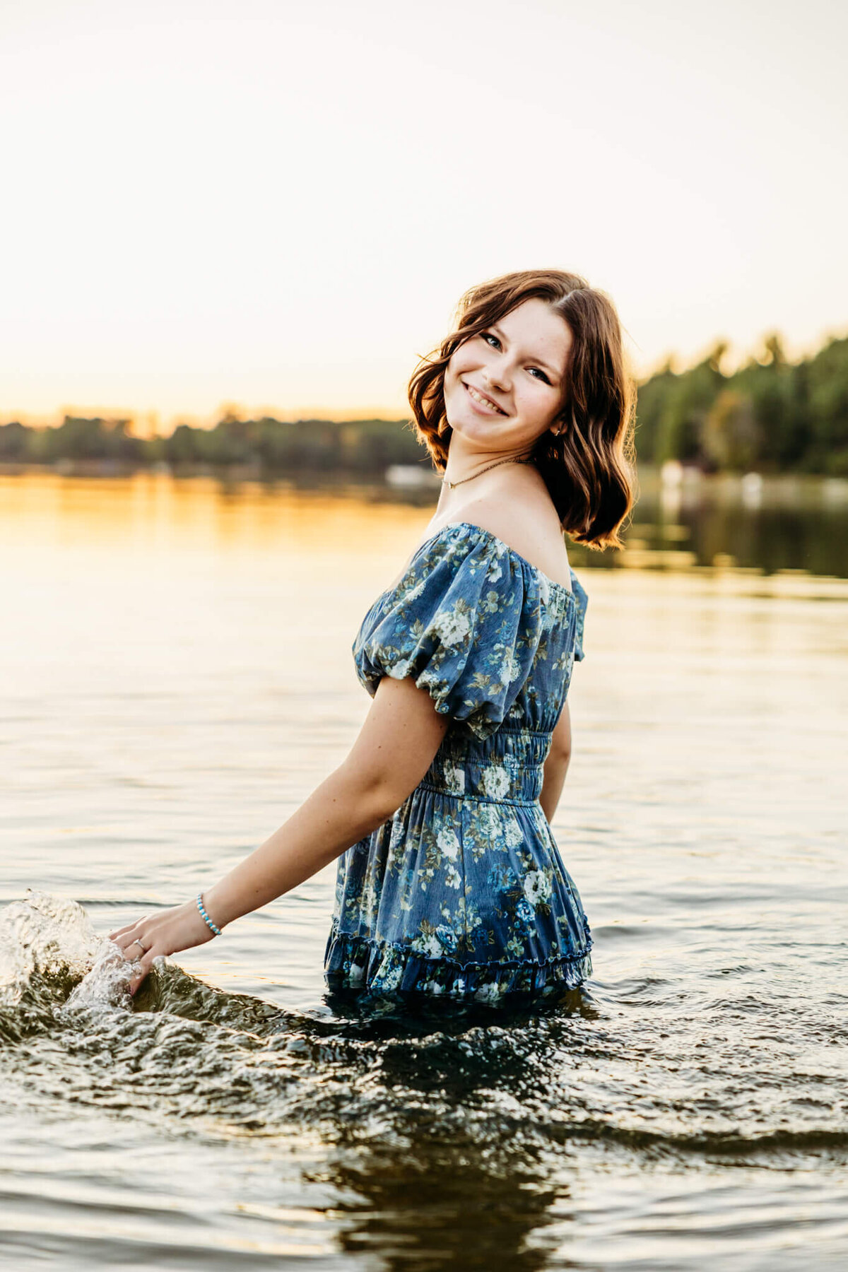 beautiful Appleton high school senior in a blue floral dress standing in the water up to her waist and brushing the surface with her hand as the sun sets behind her in a lake near Appleton.