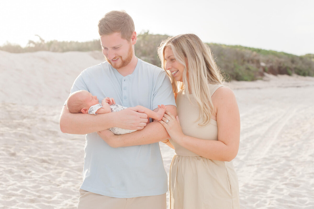 New parents smile down  at newborn on the beach with the sunsetting behind them