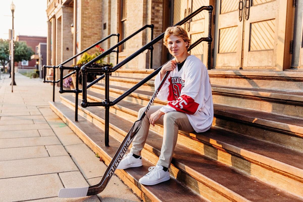 young man sitting on brown steps and resting his hockey stick on his shoulder as he looks over for his senior pictures.