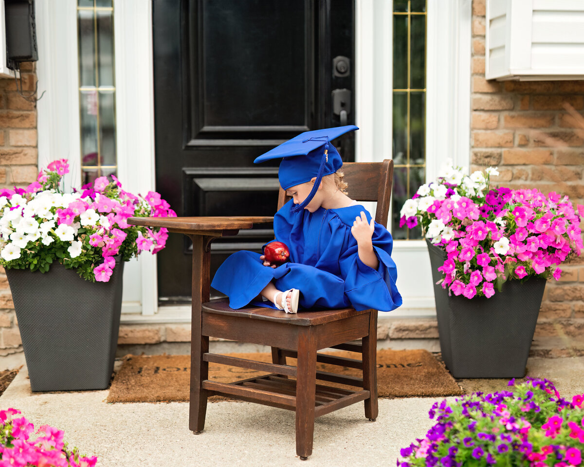 Quiet milestone moment of a little girl in graduation attire studying an apple—symbolizing knowledge, growth, and grace.