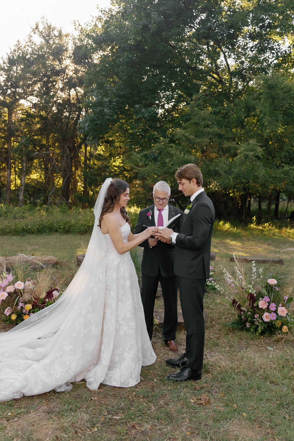 Outdoor wedding ceremony in Arkansas with the couple exchanging rings surrounded by lush, organic floral arrangements in blush, mauve, and ivory designed by a fine-art wedding florist.