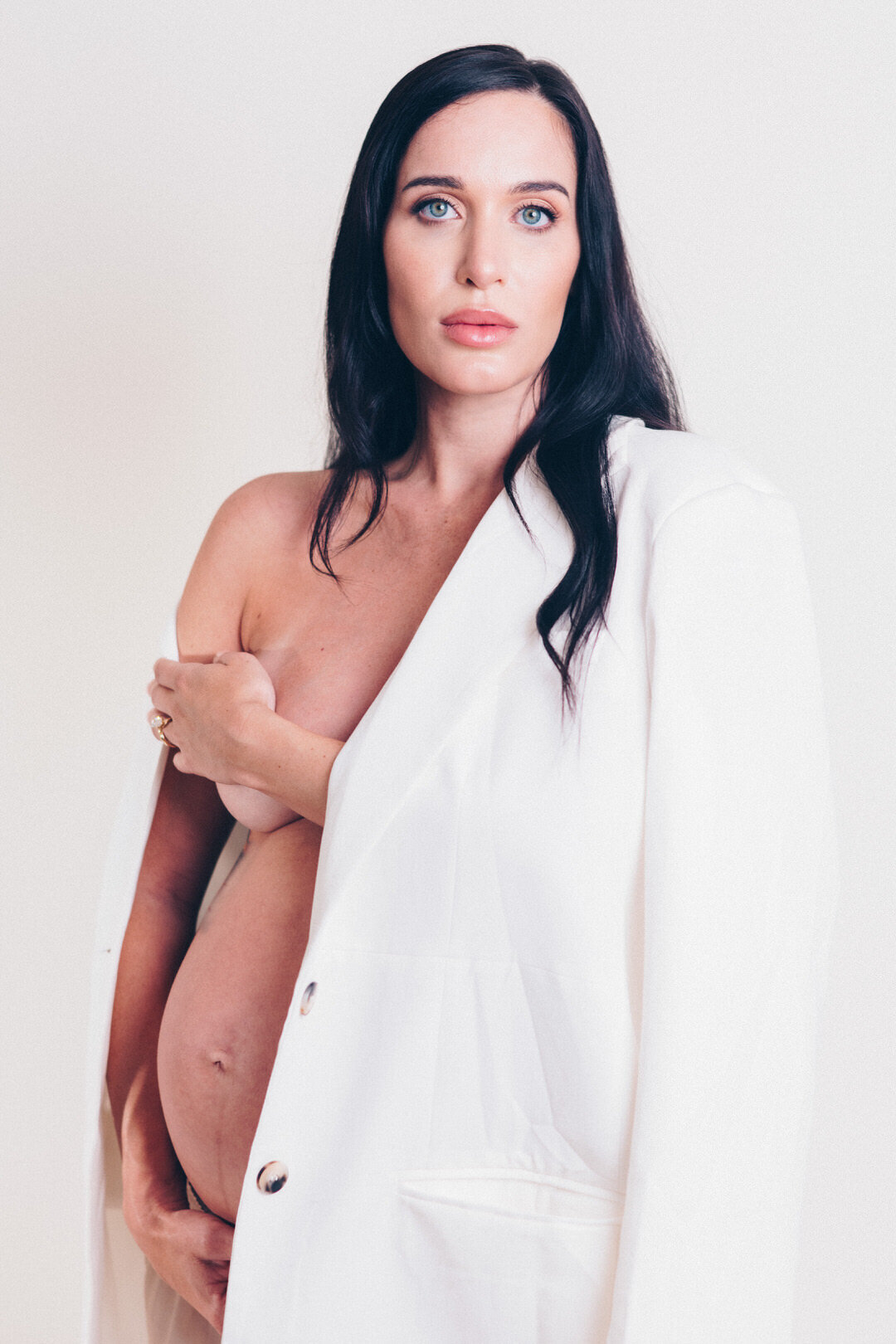 dark haired model with white blazer hanging off shoulder and exposing pregnant belly