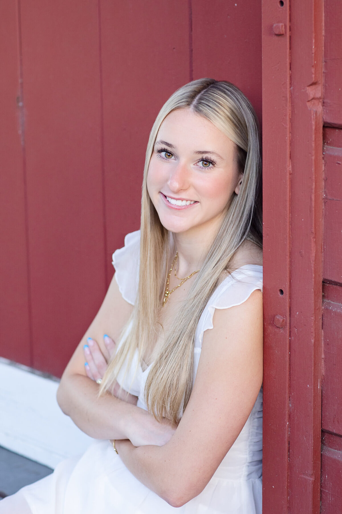 A person with long blonde hair smiles while leaning against a red wall. They are wearing a white dress and have their arms crossed. The background features a wooden texture.