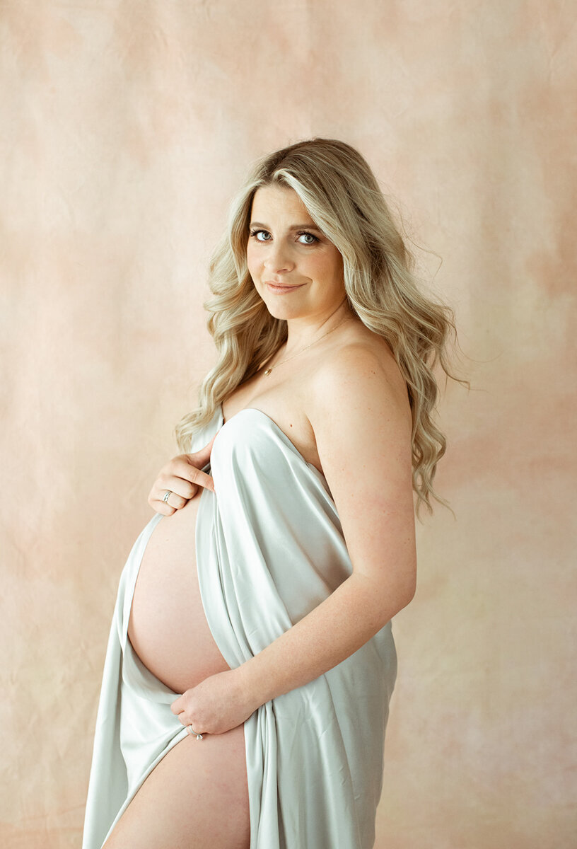 blonde pregnant mom is draped in grey silk fabric standing in front of a blush backdrop