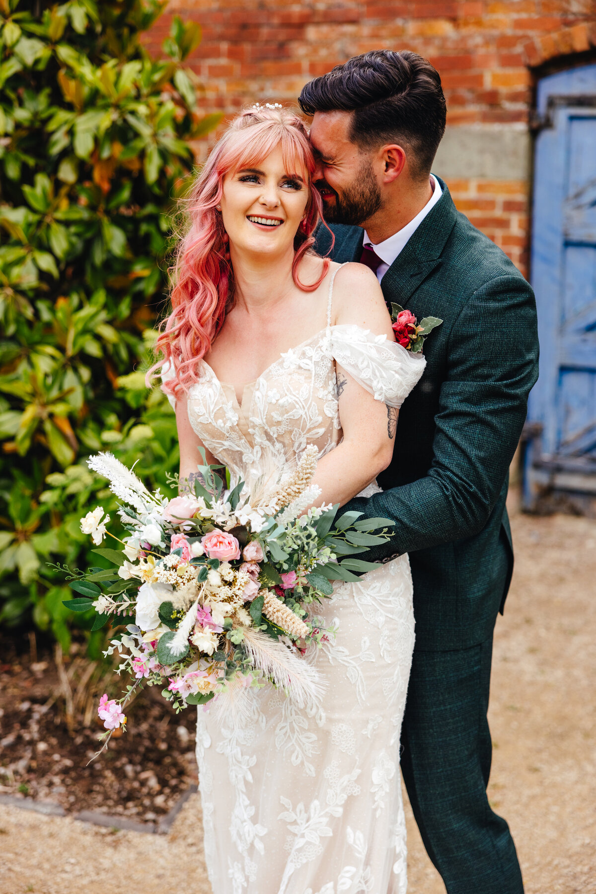 Pink-haired bride being hugged by groom from behind at their wedding at Thorpe Gardens, surrounded by lush greenery.