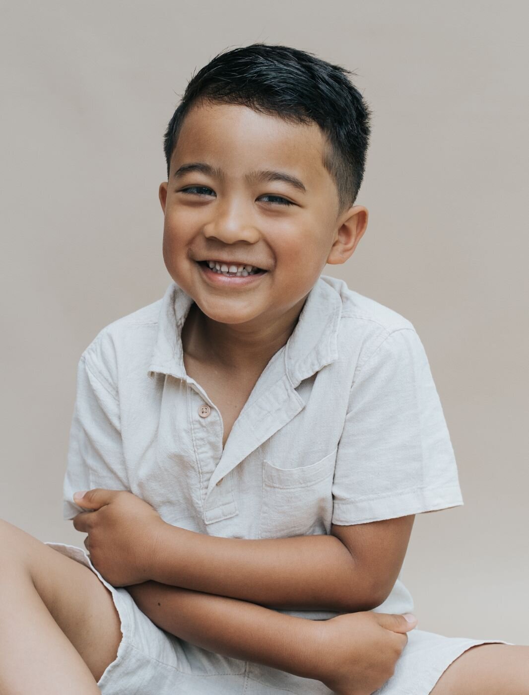 A close-up color portrait of a young boy with a joyful smile, looking directly at the camera. He is sitting with his arms crossed over his chest and is wearing a light-colored polo shirt and shorts. The background is a solid, warm tan color.